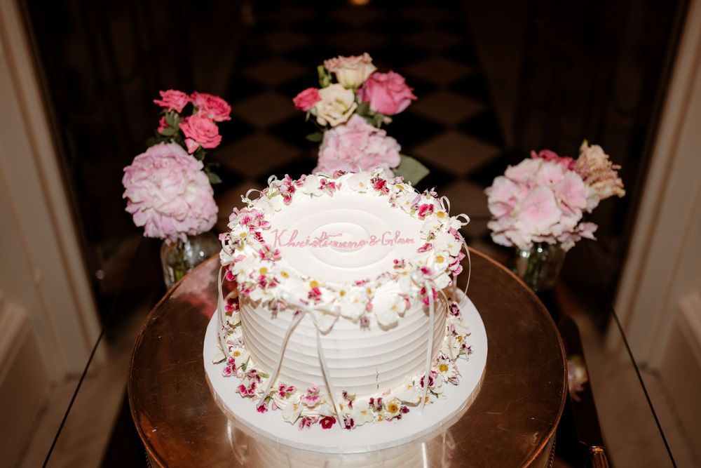 White wedding cake decorated with pink flowers and rose petals on a rustic wooden table at a reception.