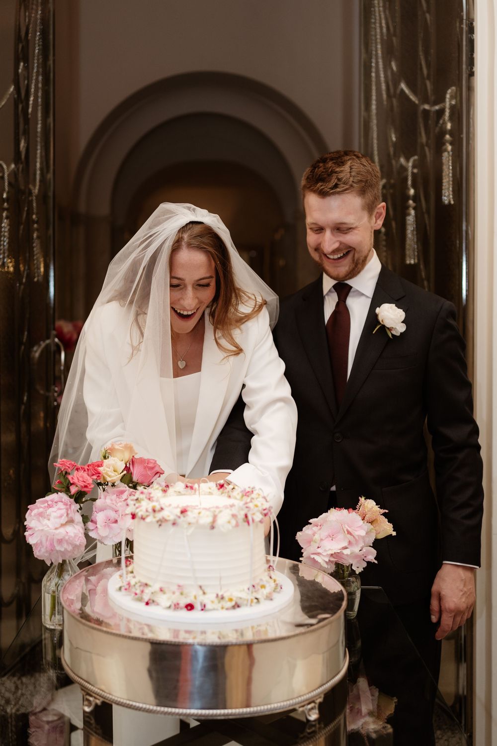 Bride and groom share a joyful moment while cutting their pink-frosted wedding cake adorned with fresh flowers.