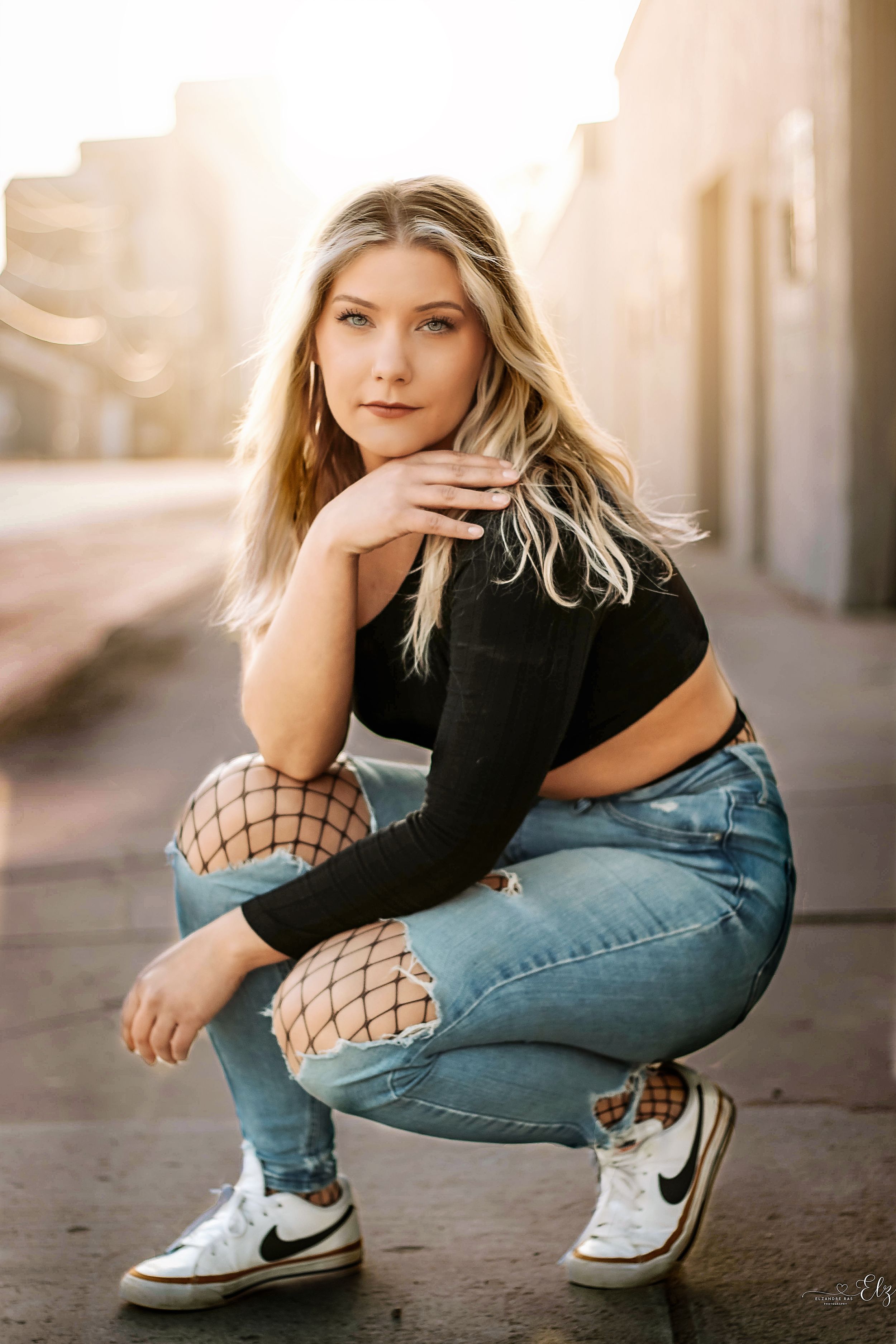 Woman in black top and jeans crouches down while wearing fishnet stockings and Converse sneakers in urban setting in Williston, North Dakota
