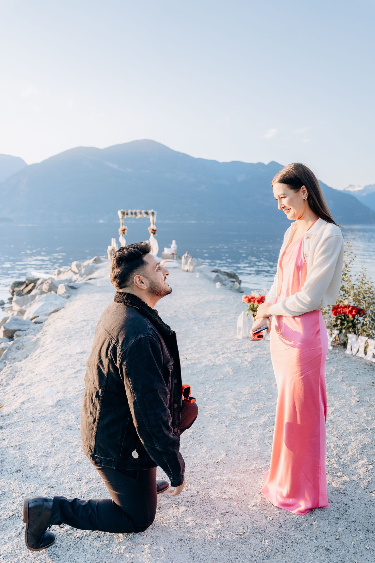 A marriage proposal scene unfolds on a rocky beach with mountains in the background as someone kneels with a ring.