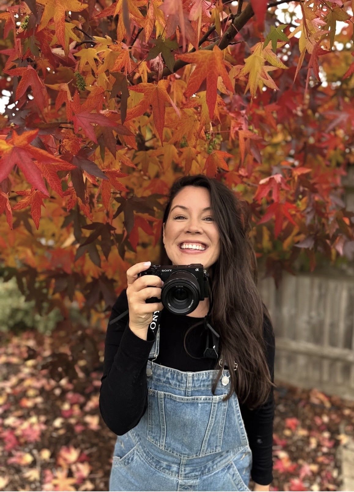 A photographer with a camera poses under vibrant red and orange autumn leaves. Photography in Buckinghamshire.