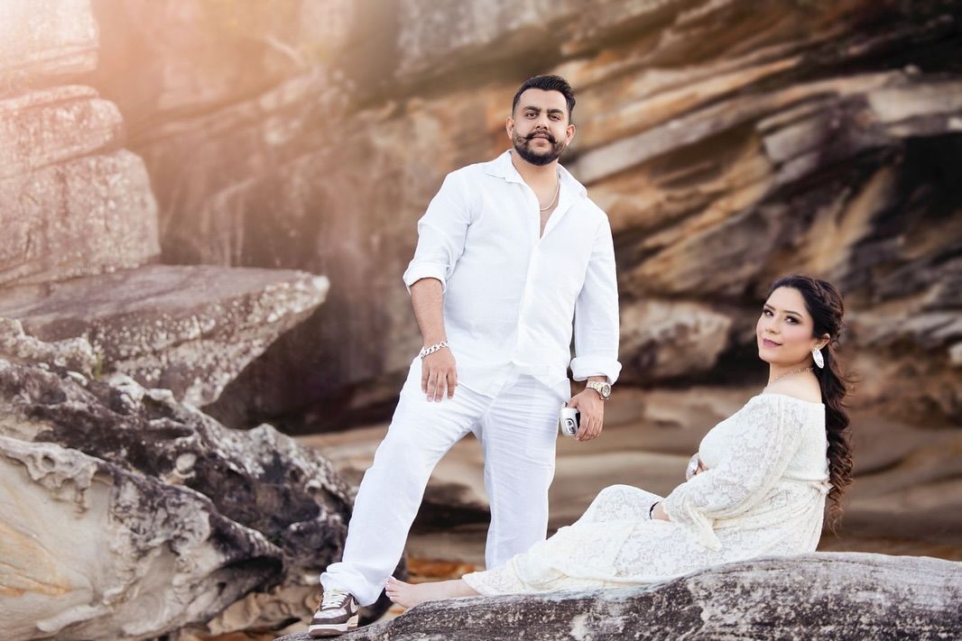 A couple in white outfits poses together on rocky terrain during a romantic photoshoot at sunset.