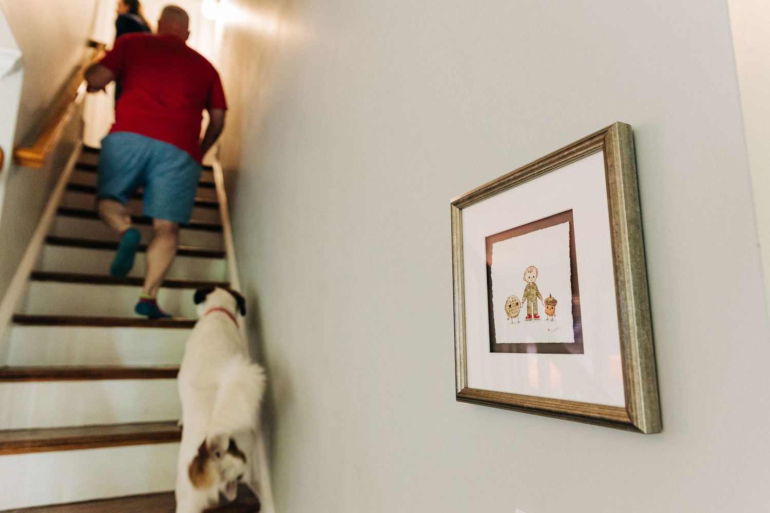 A person walks up wooden stairs with a small dog following behind, while a framed artwork hangs on the white wall.