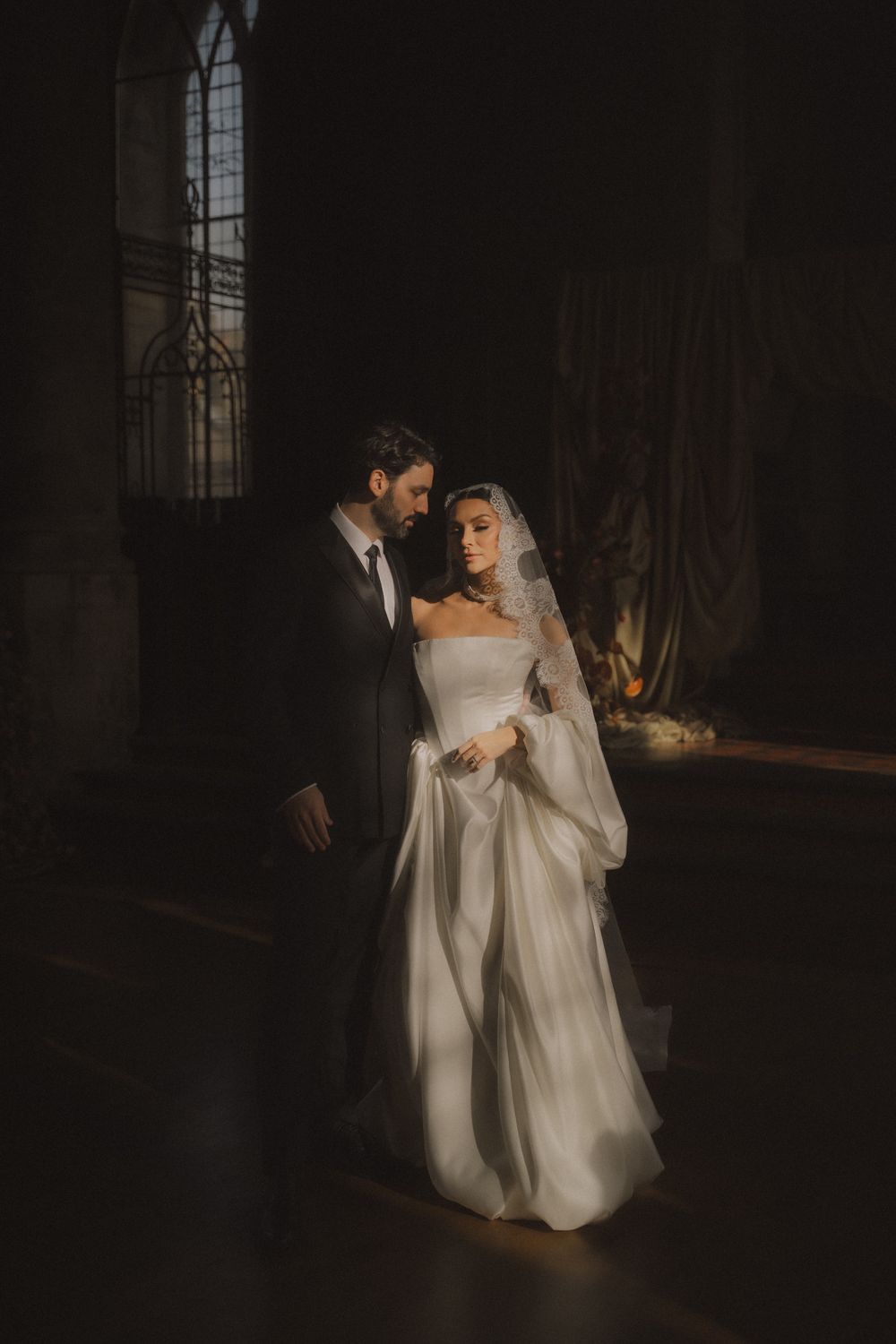 A romantic wedding couple dances together in a dimly lit church with dramatic shadows and light streaming through windows.