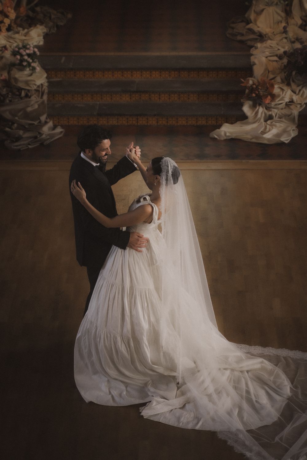A romantic wedding couple embraces in an intimate dance pose against a rustic dark background with dramatic lighting.