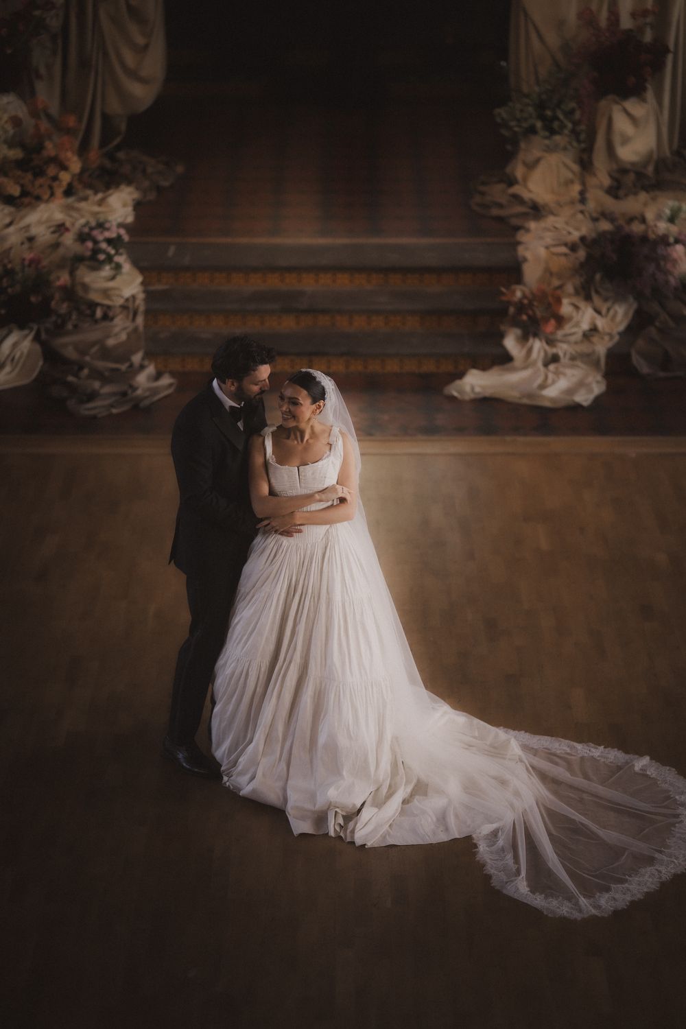 A romantic wedding couple dances in an elegant dark venue with flowing white dress and long veil trailing behind them.