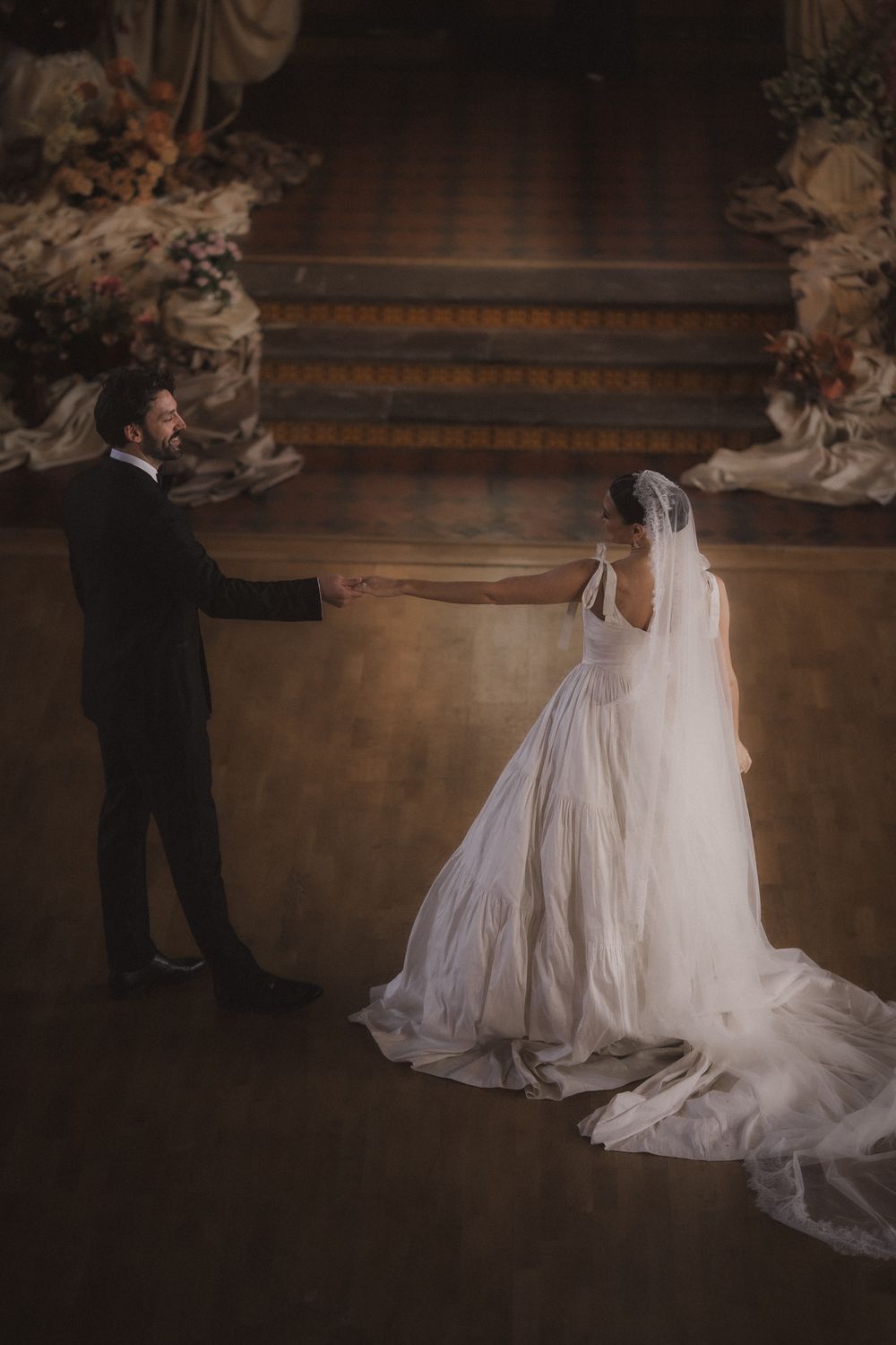 A romantic wedding couple dances together in an elegant ballroom with ornate architectural details and soft lighting.