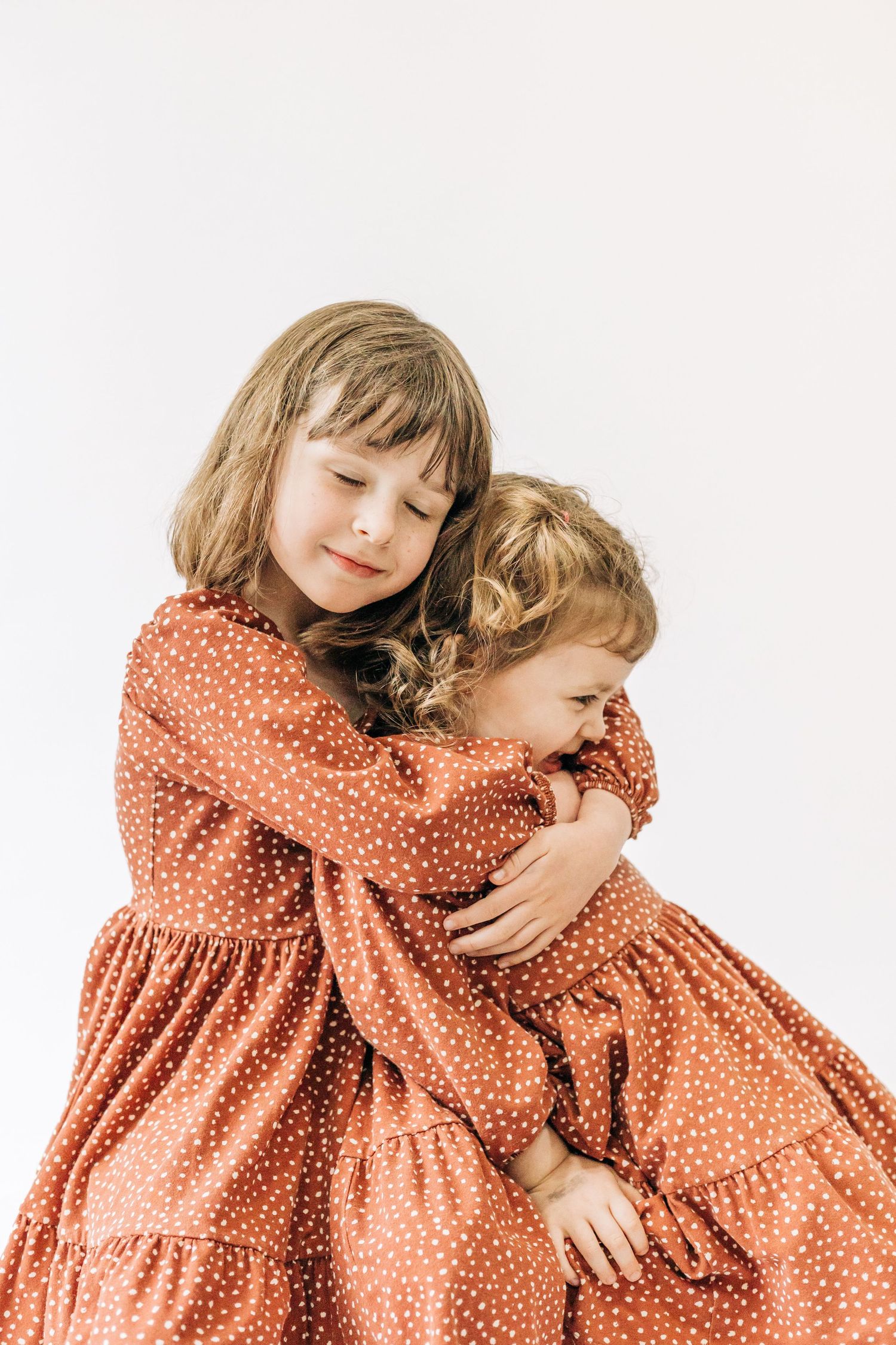 Two children in matching polka dot orange dresses embrace each other against a white background.