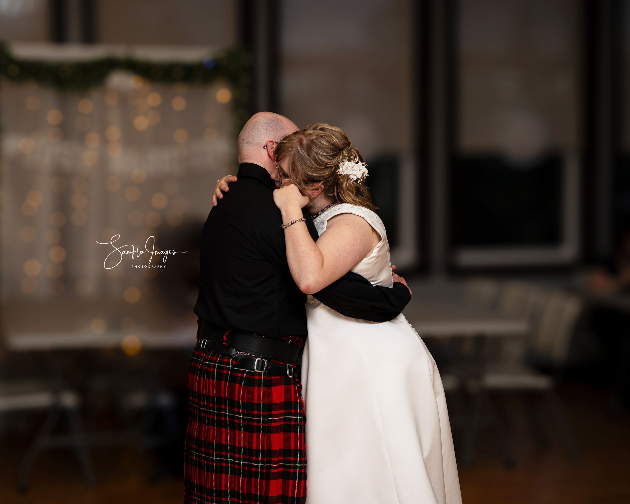 A couple embraces in a romantic dance pose at a wedding reception with warm lighting and festive decor.