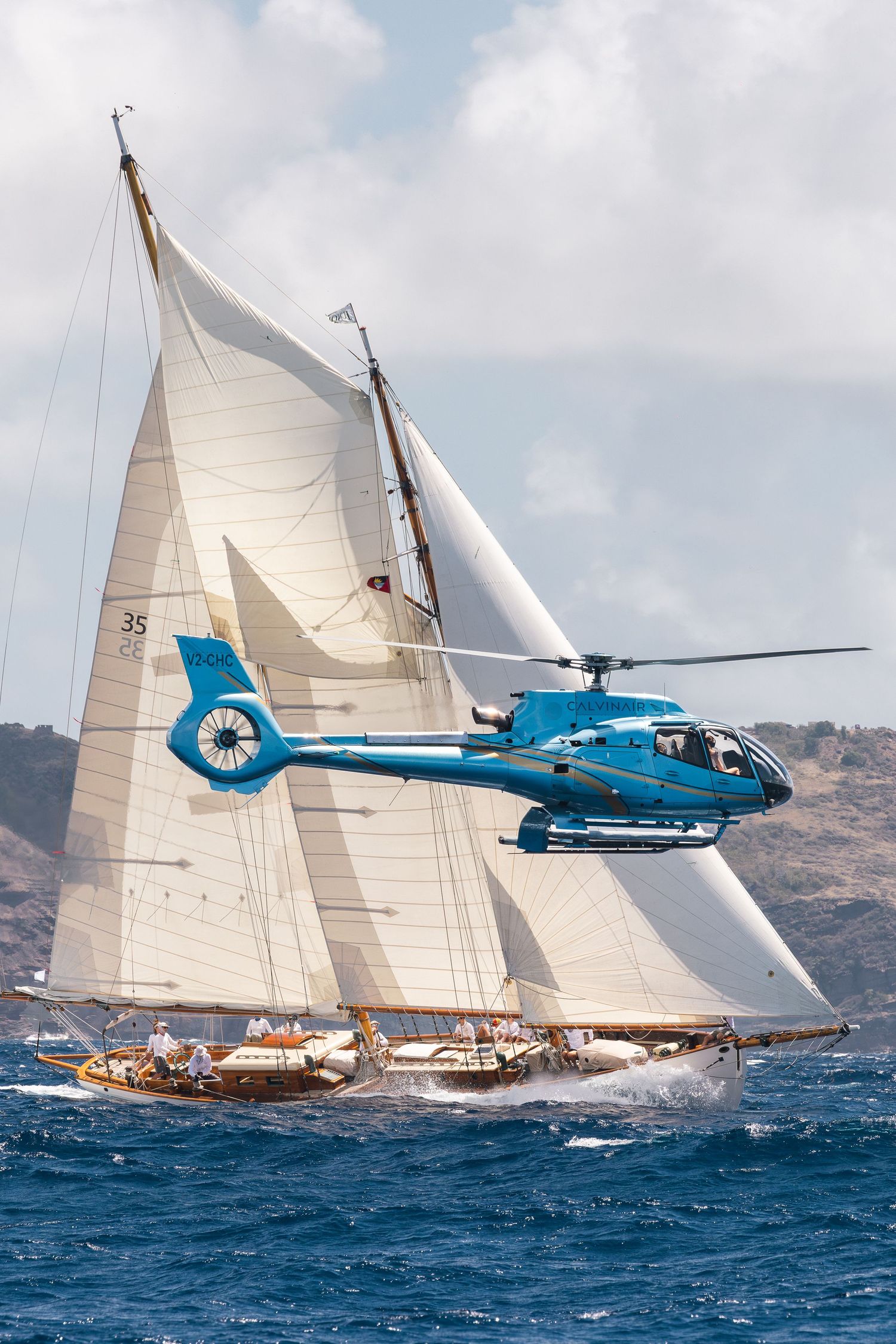 Blue helicopter flying alongside a classic tall ship with white sails racing on blue ocean waters.