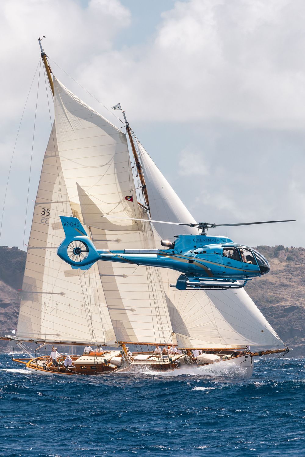 Blue helicopter flying alongside a classic tall ship with white sails racing on blue ocean waters.