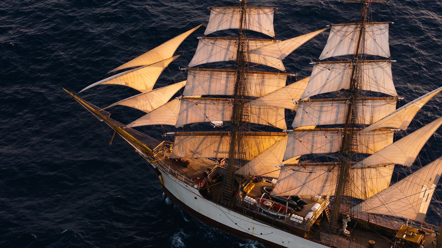 Aerial view of a tall ship with full white sails navigating the deep blue ocean at golden hour.