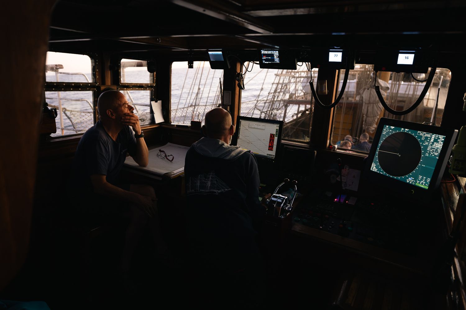 Two crew members navigate a dimly lit ship bridge with radar screens and controls at sea.