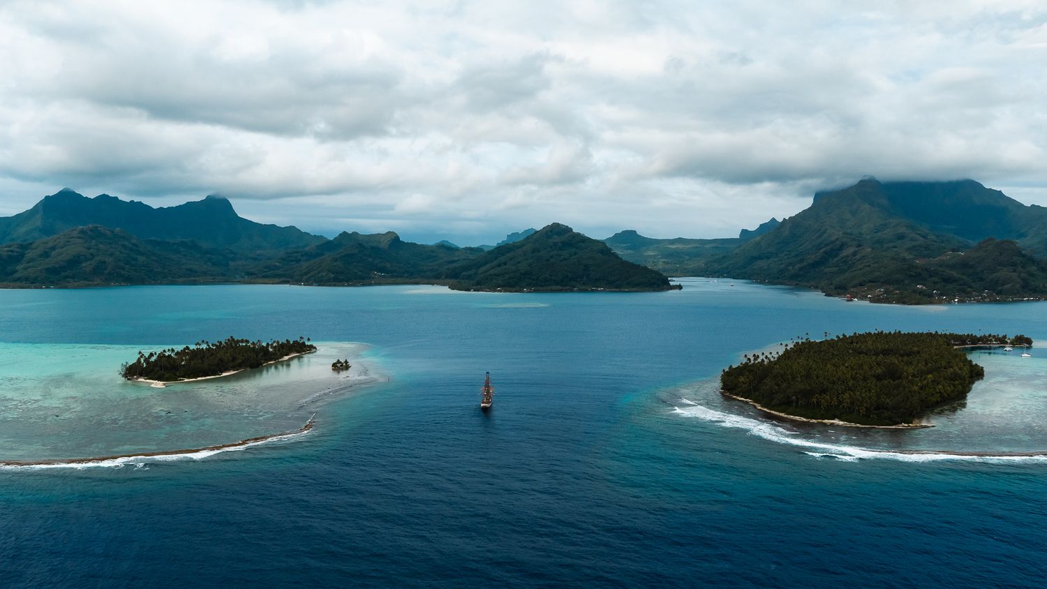 Aerial view of lush tropical islands surrounded by turquoise waters with dramatic mountain backdrop under cloudy skies.