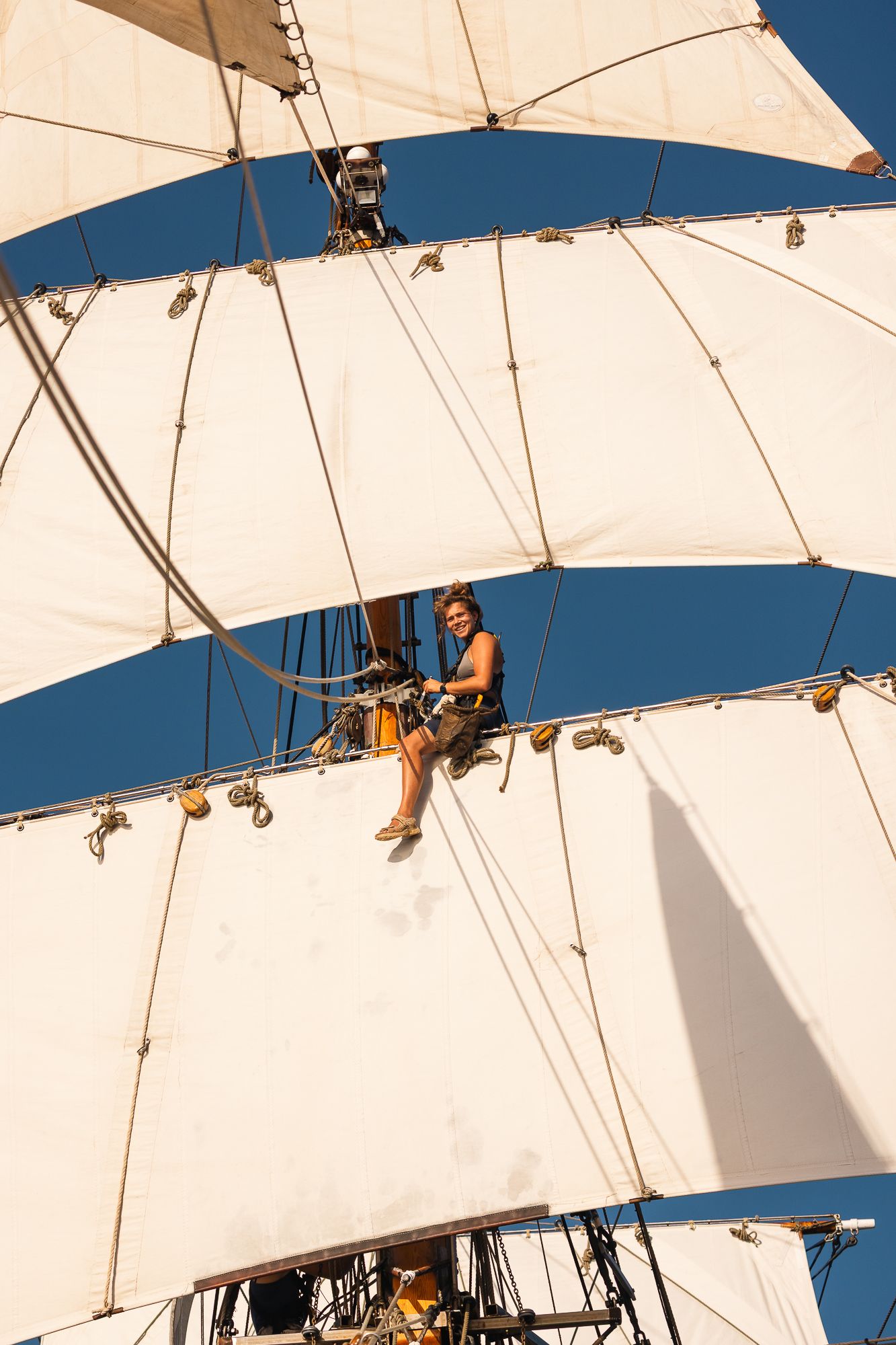 Shirtless sailor climbing tall ship mast rigging between white sails against clear blue sky.