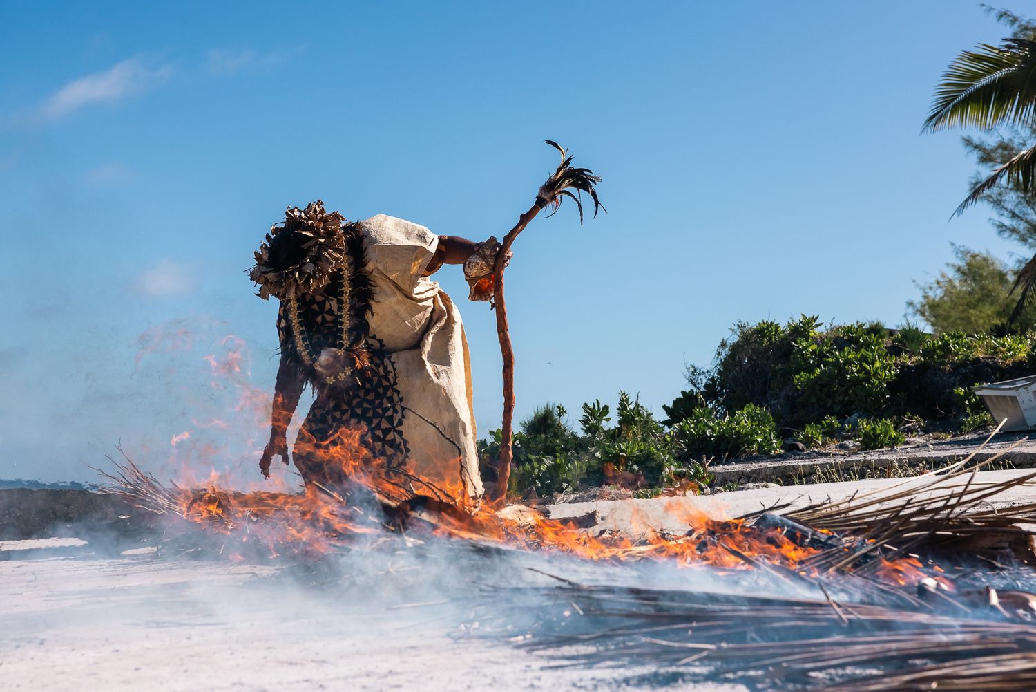 A costumed figure performing a fire ritual dance on a beach with palm trees under a blue sky.