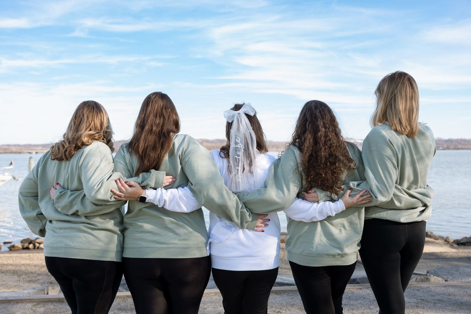 Bride surrounded by bridesmaids wearing matching sweatshirts — candid wedding celebration portrait