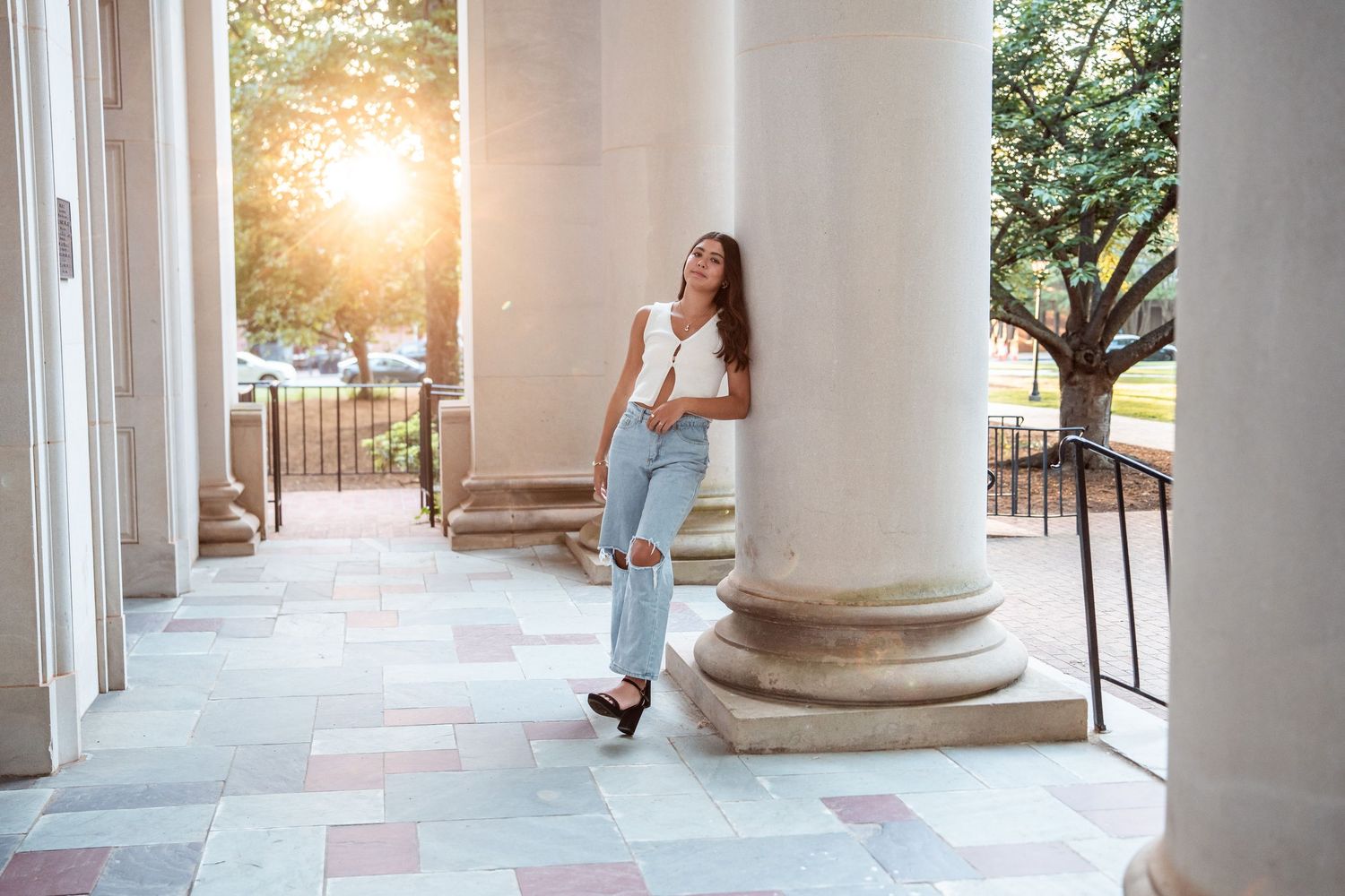 Young woman in casual attire leans against white column on porch with sunlight filtering through trees in background.