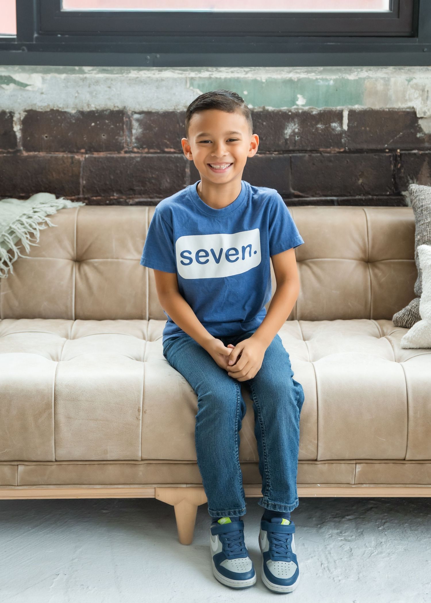 Young person in a blue "seven" t-shirt and jeans sits smiling on a beige tufted couch against a rustic brick wall.