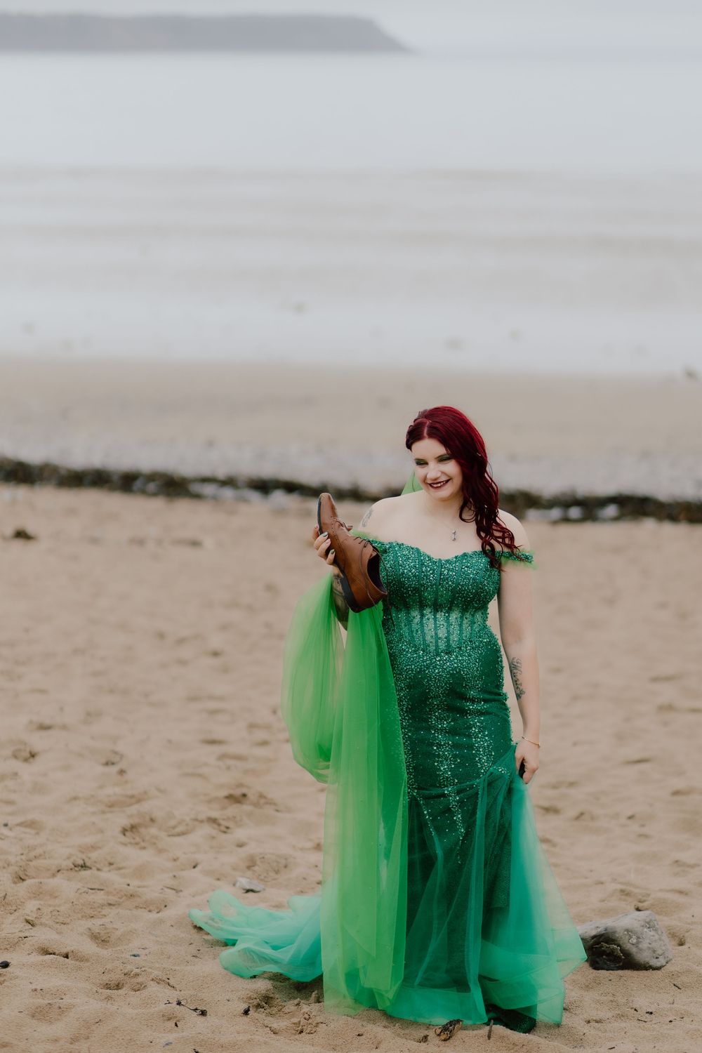 A woman in a sparkly emerald green gown with flowing tulle cape stands on a misty beach.