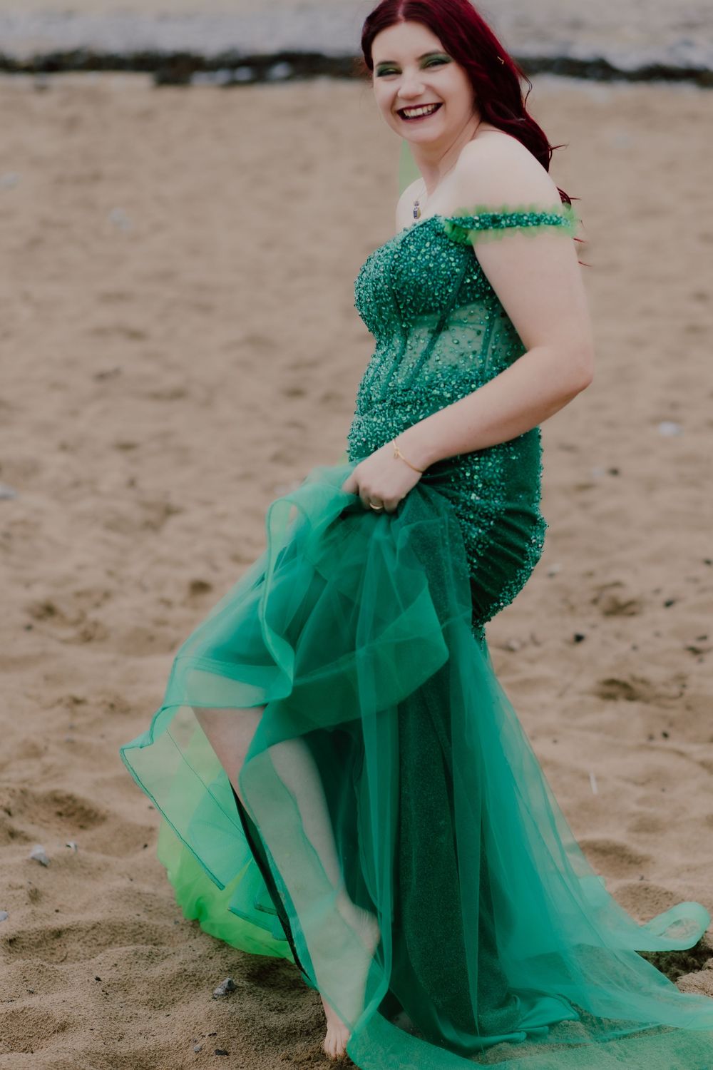 A person in a sparkly emerald green tulle gown poses on a sandy beach.
