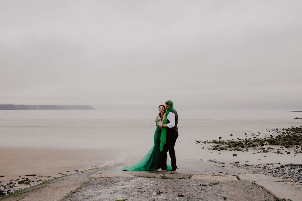 A couple in formal attire embrace on a misty beach, with the woman wearing a flowing emerald green dress.
