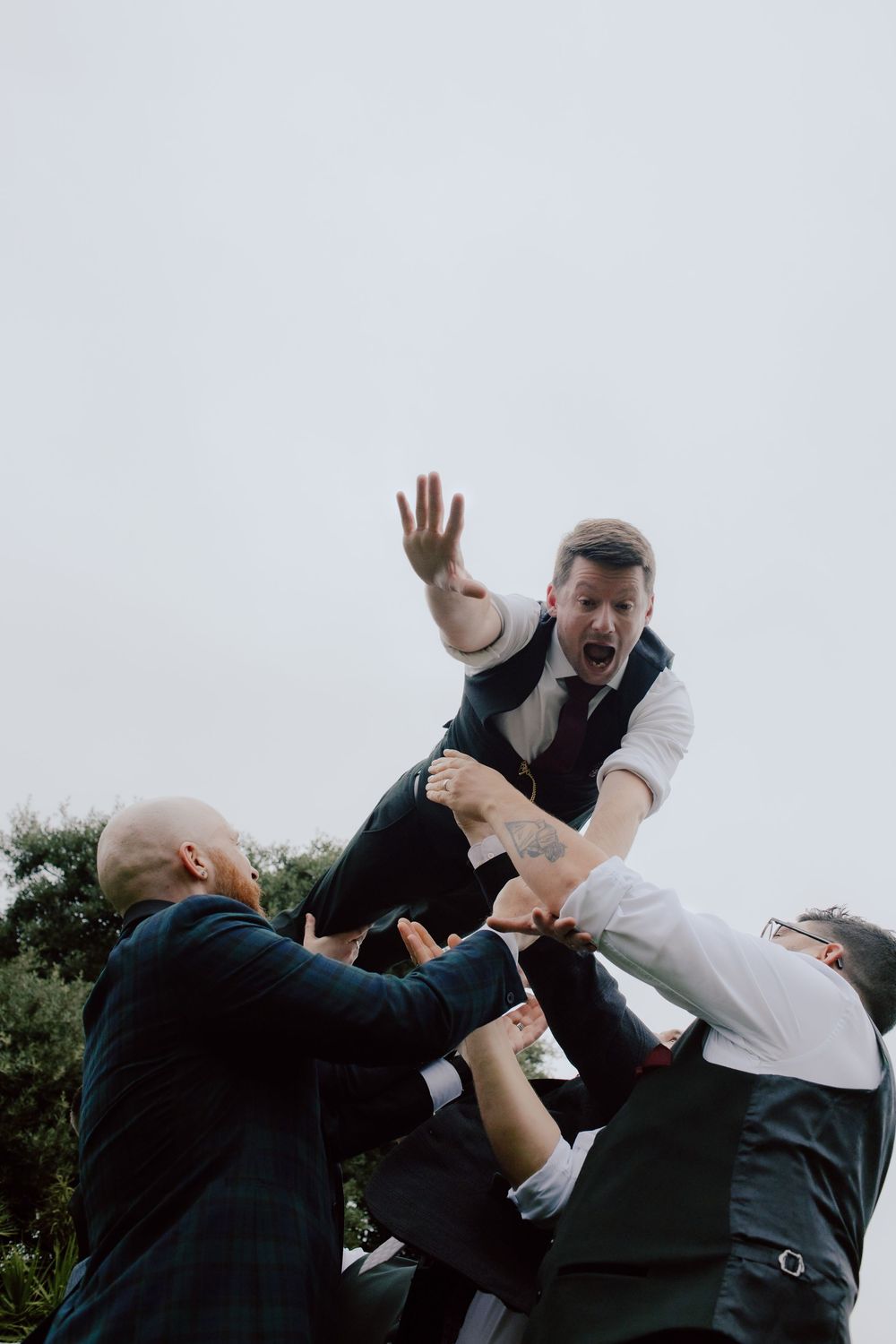 Groomsmen playfully lift and toss someone in formal attire against a cloudy sky during a wedding celebration.