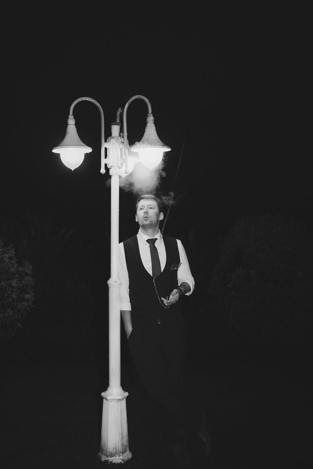 Black and white photo of a person posing mysteriously near vintage street lamps at night.