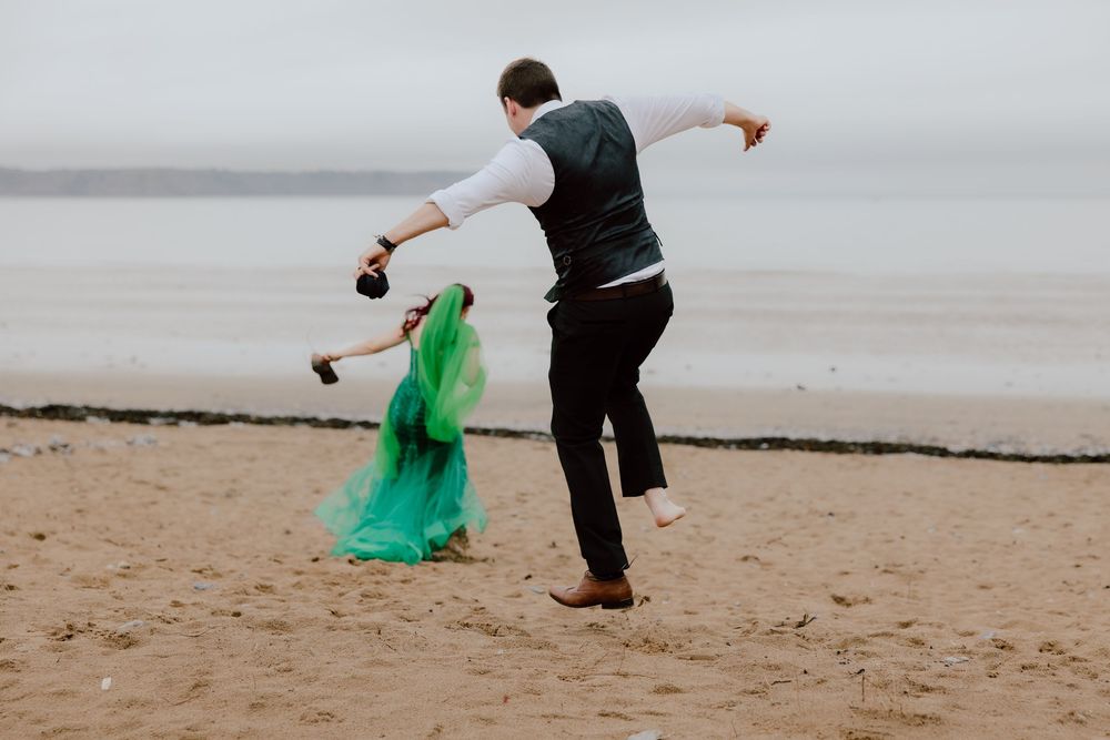 A person in dark clothing releases a green fabric that flows in the wind along a misty beach.