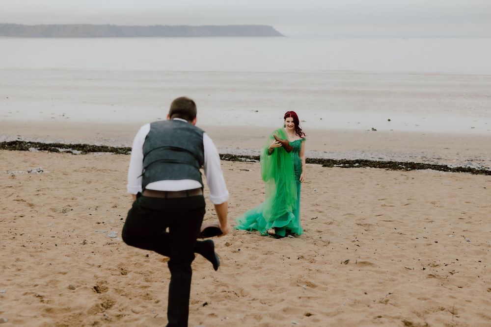 Two people interact on a misty beach, with one wearing a flowing emerald green dress while another poses in dark attire.