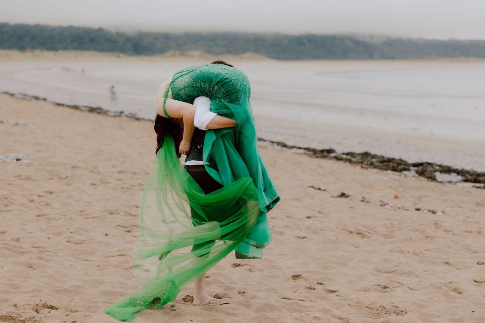 A figure in flowing emerald green fabric dances on a misty beach shoreline.