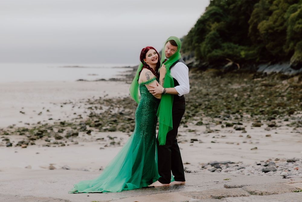 A couple in elegant green formal attire embraces on a rocky beach during an overcast day.