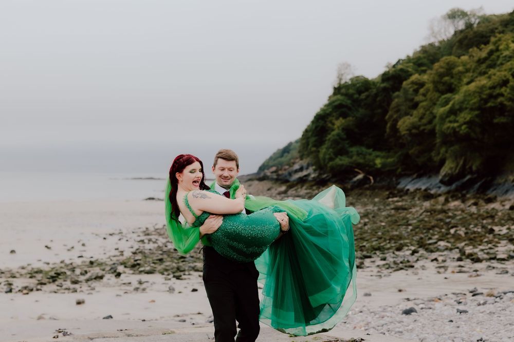 A couple in emerald green attire dances together on a rocky beach with dramatic coastal cliffs in the background.