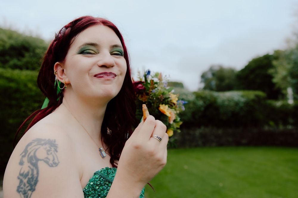 Someone with burgundy hair wearing a green dress holds a small bouquet of flowers outdoors in a garden setting.