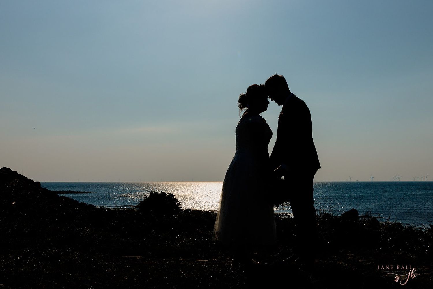 A silhouetted couple stands close together against a sunset ocean backdrop creating a romantic coastal scene.