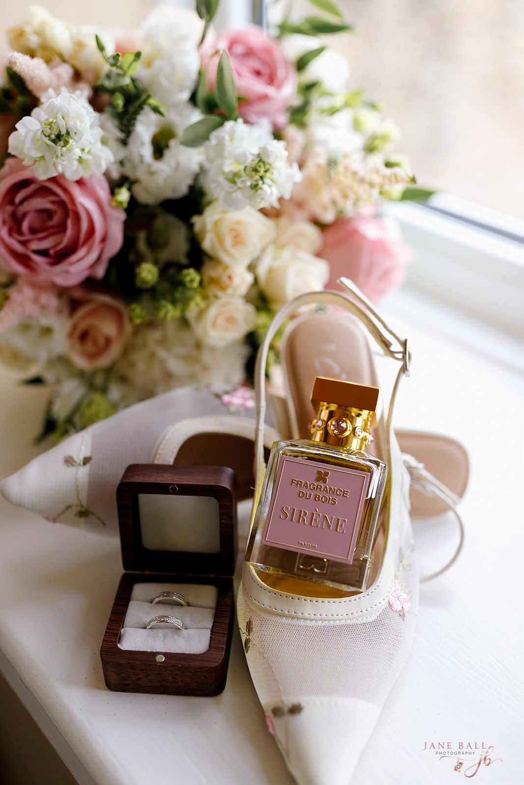 Elegant wedding details with pink roses, white flowers, perfume bottles and a ring box displayed near a window.