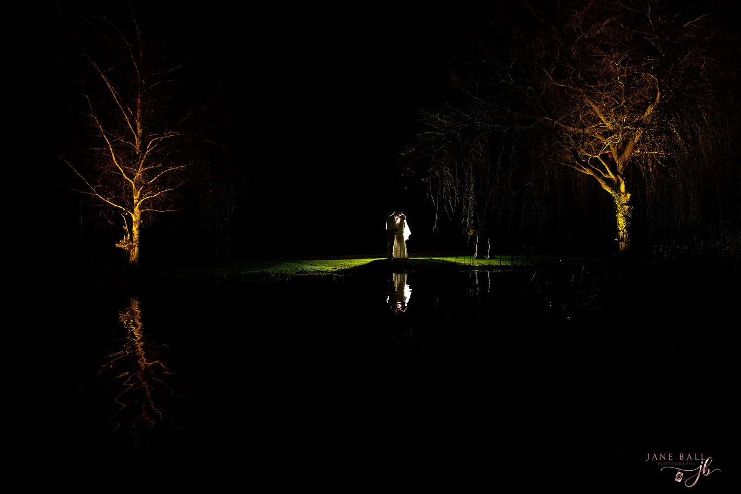 Silhouetted figure stands between illuminated trees reflecting on still water at night in dramatic lighting.