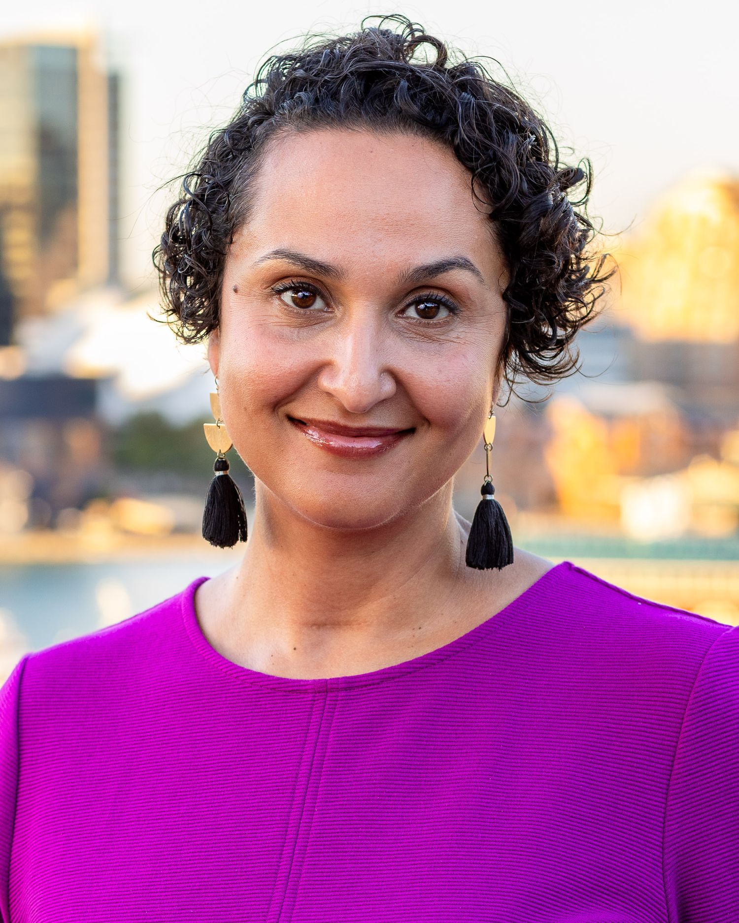 A professional woman in a vibrant magenta top smiles warmly against a soft-focus cityscape.