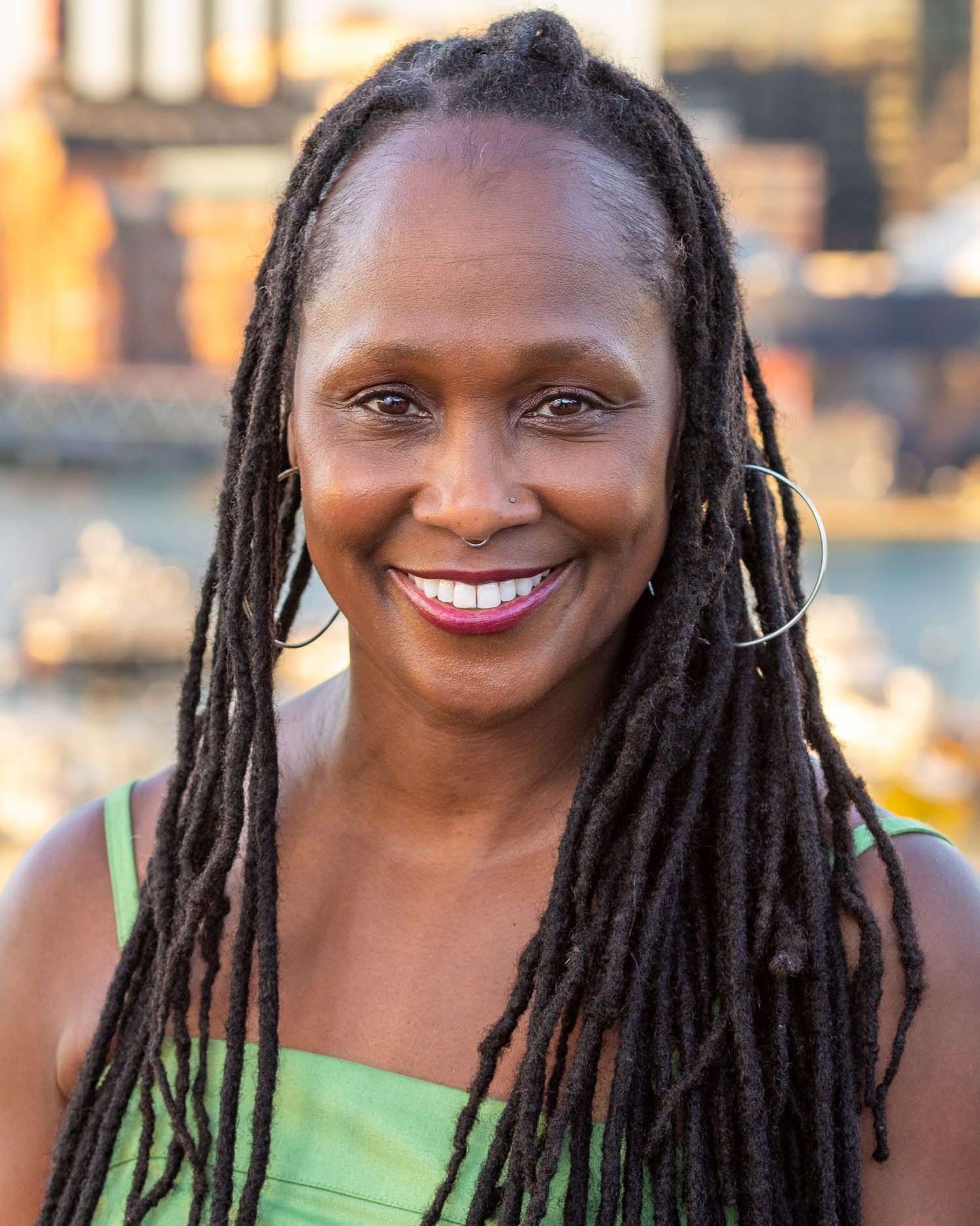 A professional woman with long dreadlocks and a bright smile wearing a green top against a blurred cityscape.