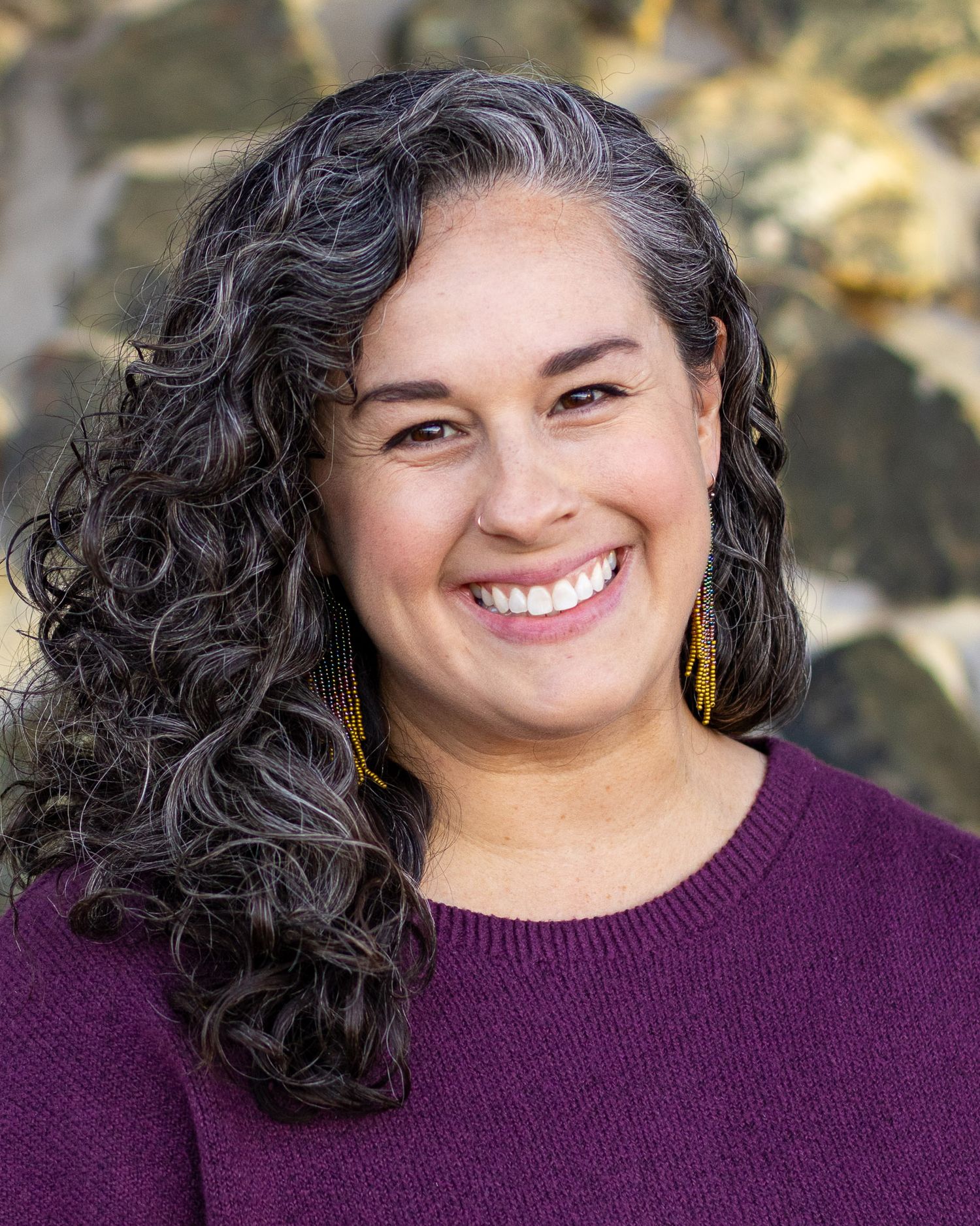 A person with dark curly hair and a burgundy sweater smiles warmly for her headshot in front of a stone wall.