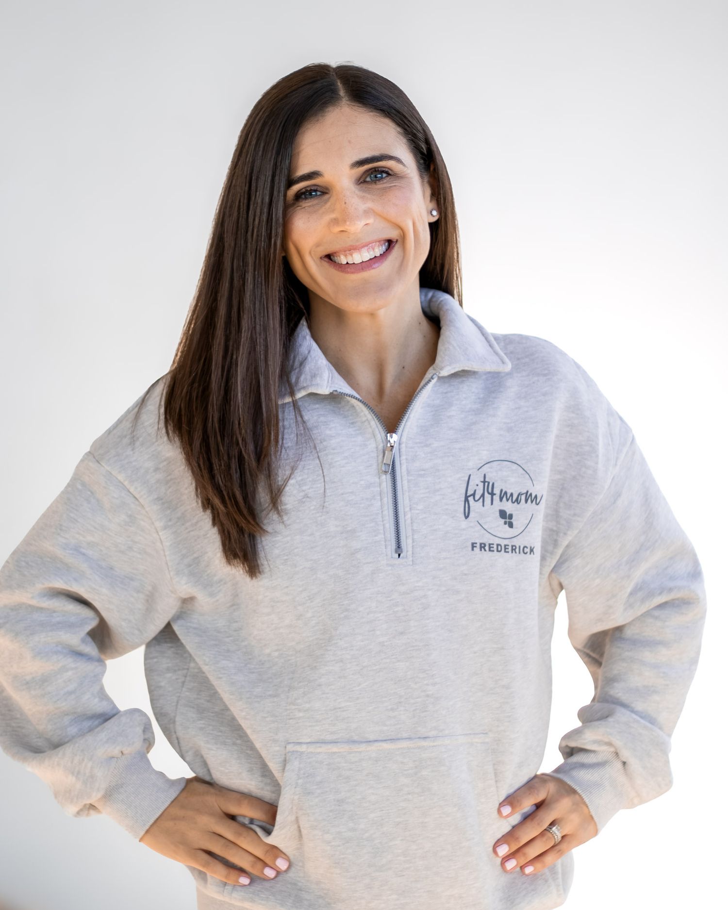 A headshot of a smiling person wearing a light gray quarter-zip pullover sweatshirt with hands on hips against a white background.