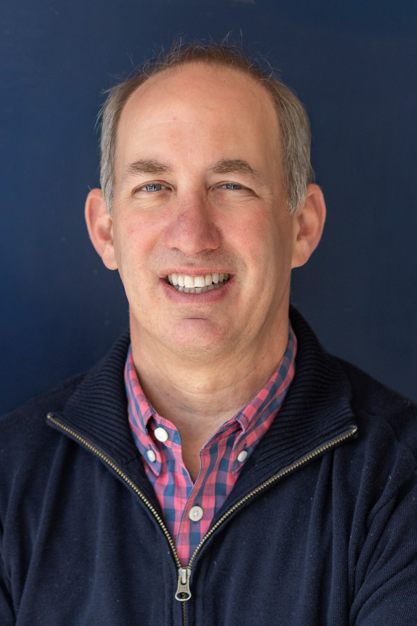 Professional headshot of a man wearing a navy vest and plaid shirt against dark background.