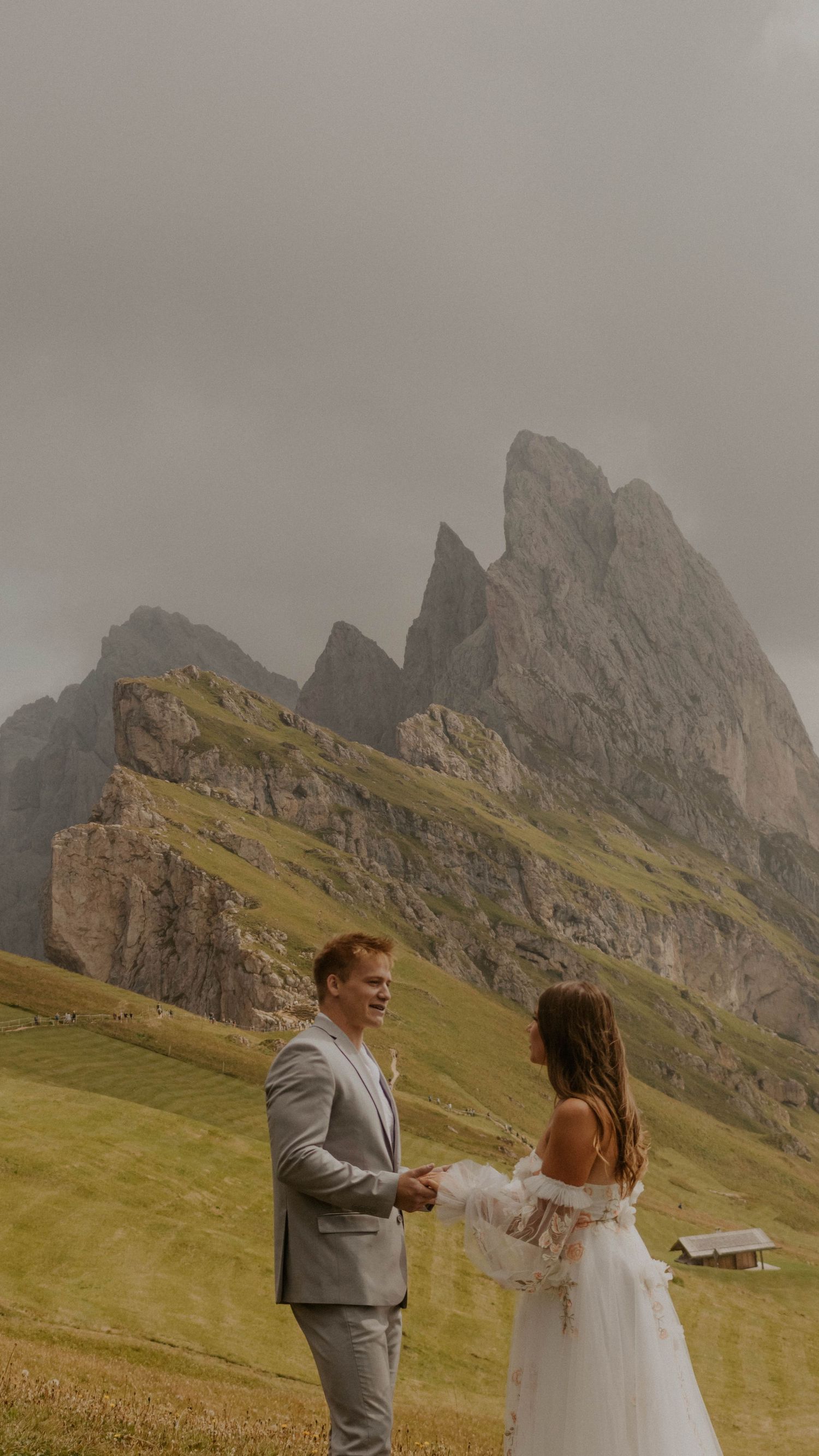 A couple in wedding attire dances together on a grassy mountainside with dramatic rocky peaks in the background.