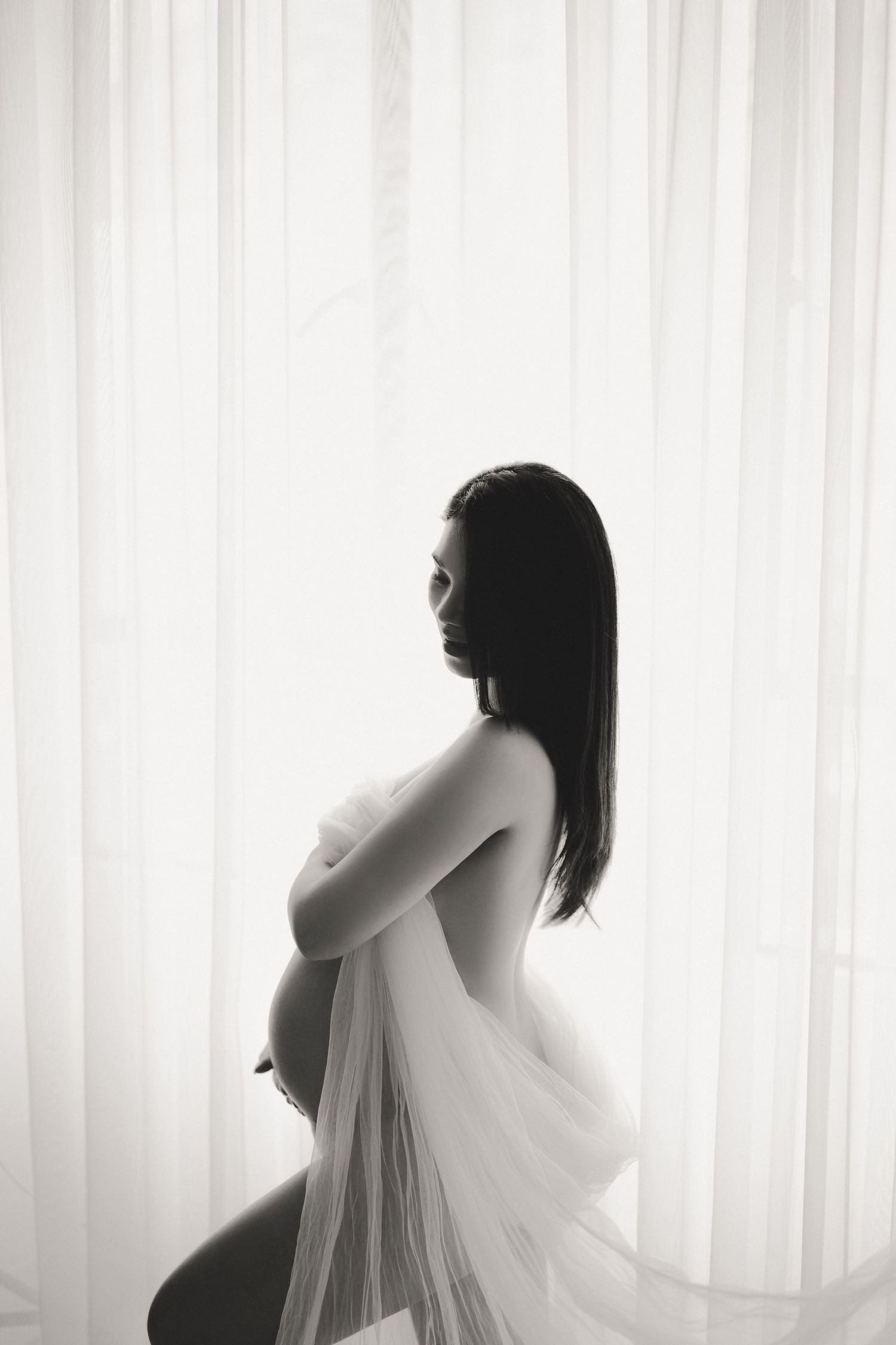Artistic black and white maternity silhouette photo with flowing sheer fabric against backlit curtains.
