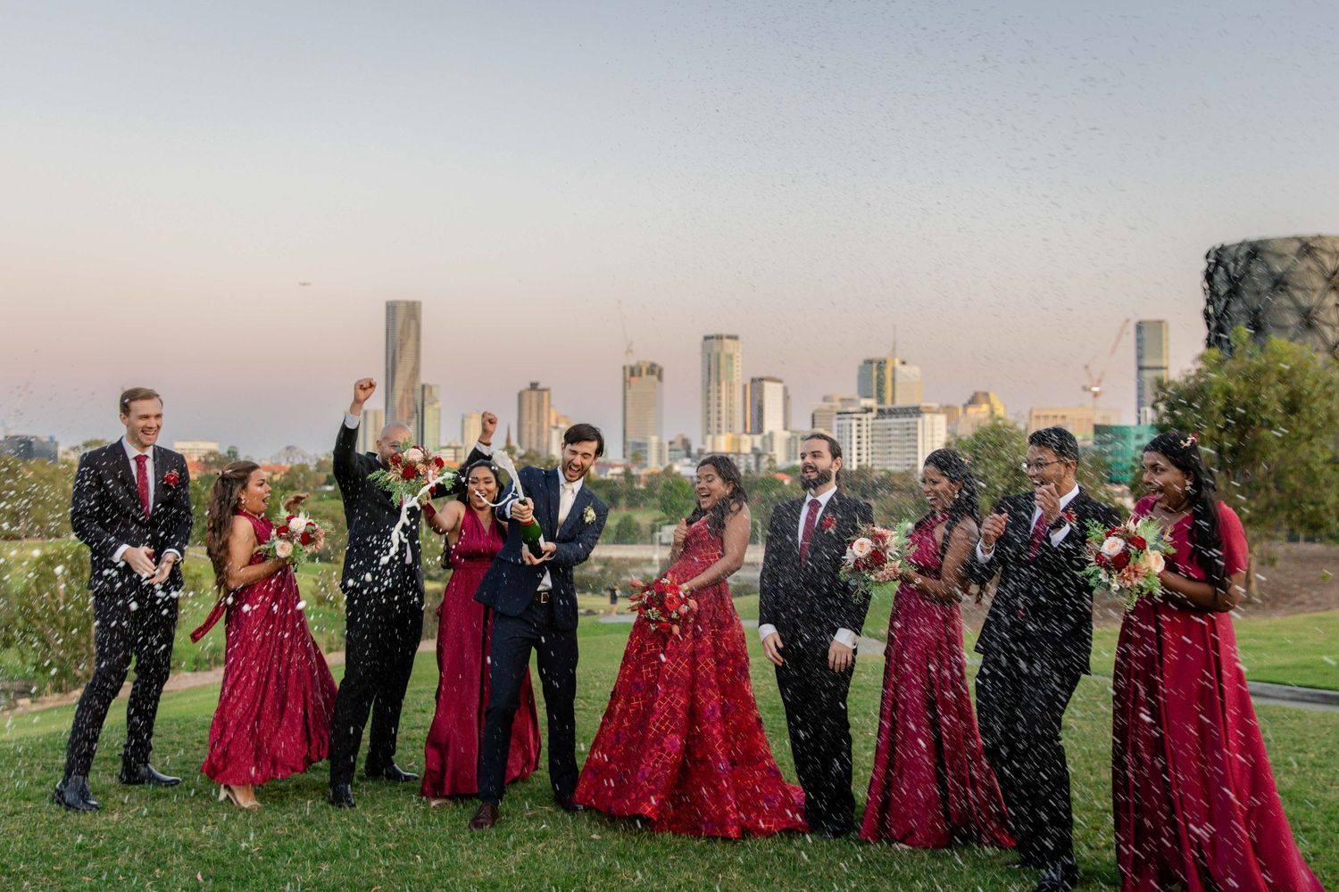 Wedding party in red gowns and suits celebrating with confetti against Melbourne city skyline at dusk.