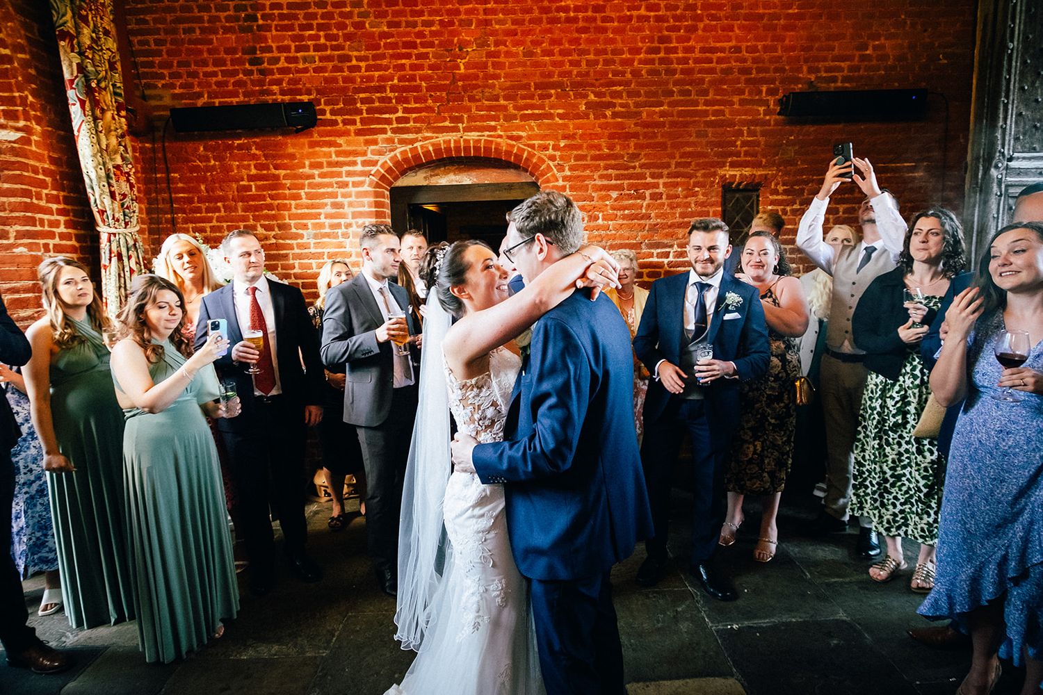 Wedding guests dance and celebrate at a reception in a rustic brick-walled venue with festive lighting.