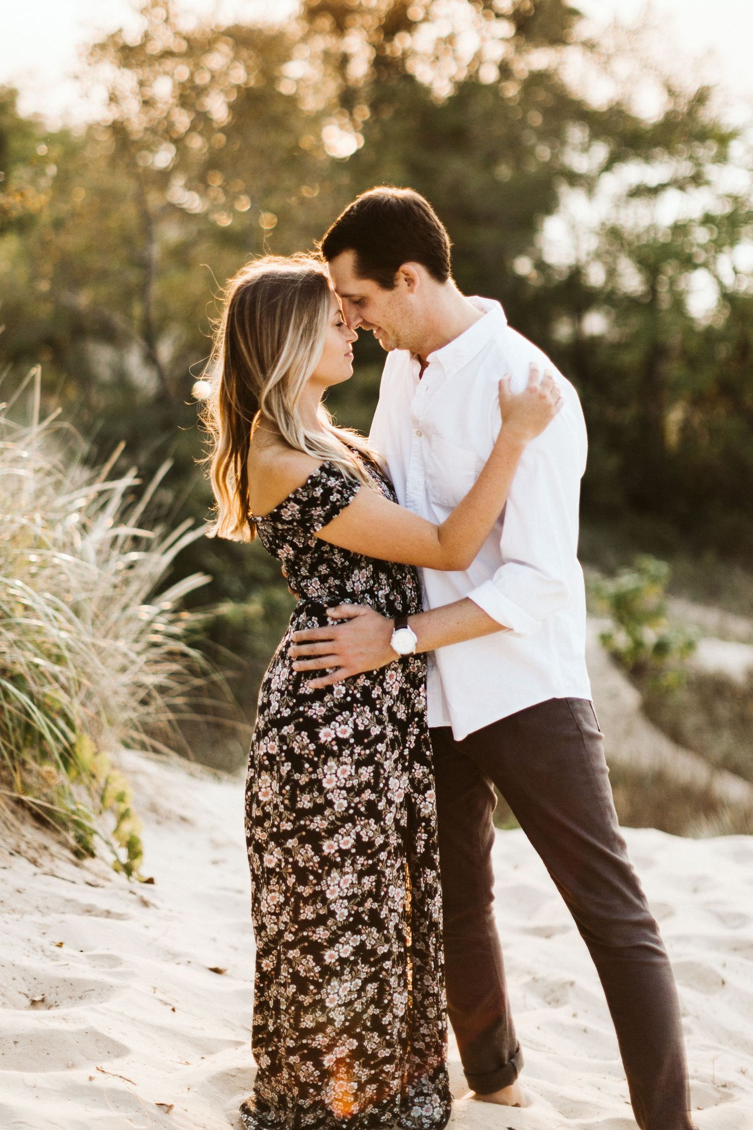Intimate couple moment on beach with floral dress and white shirt against natural backdrop.
