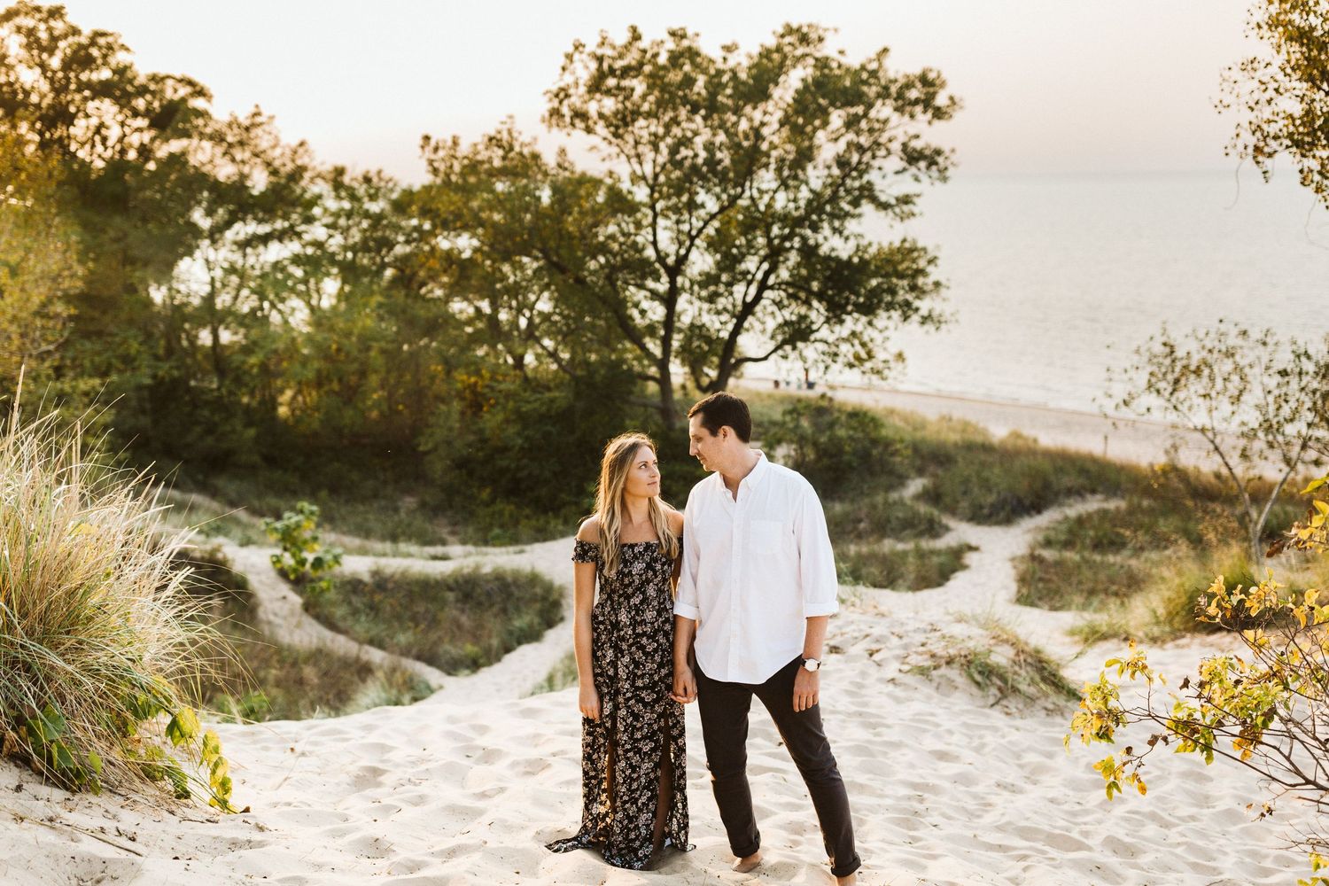 Couple walking together on a sandy beach path during golden hour sunset with trees in background.