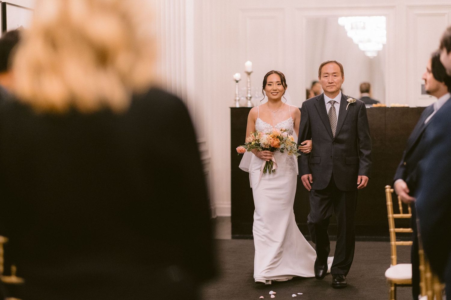A bride in a white fitted dress walks down the aisle with an escort at an indoor wedding ceremony.