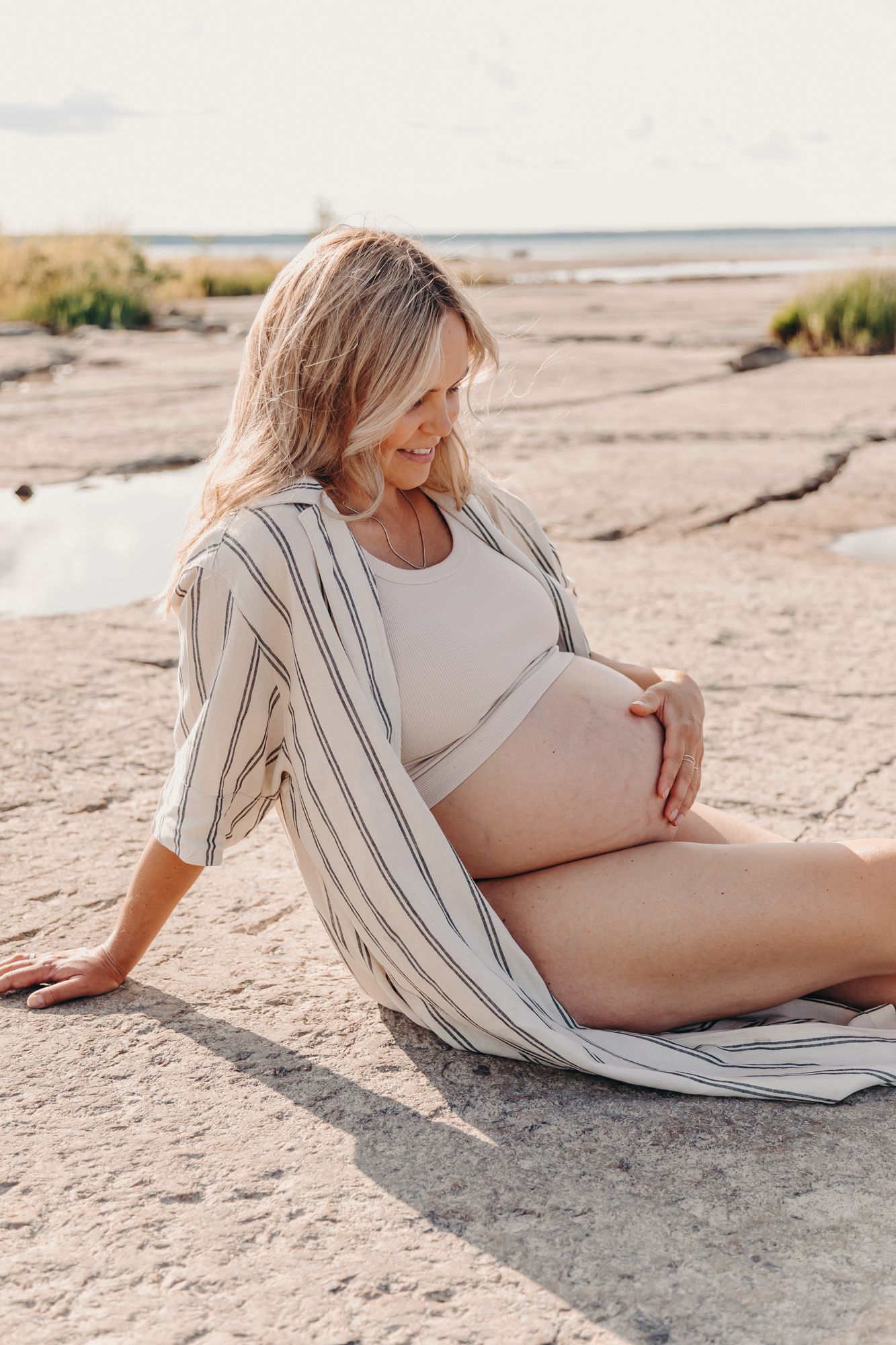 A pregnant woman in a striped cardigan relaxes on a sandy beach during sunset.